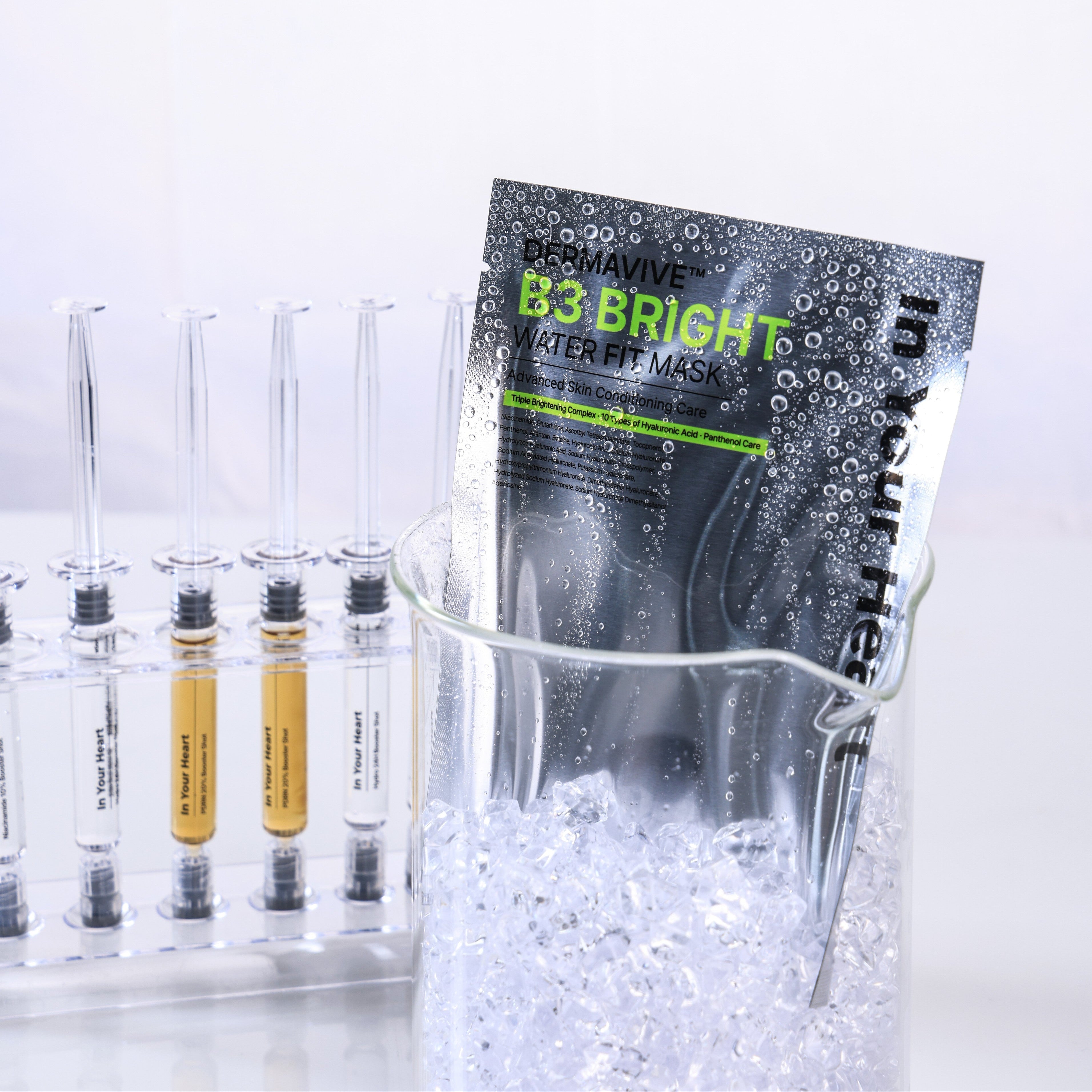 A packet of In Your Heart DermaVive B3 Bright Water Fit mask sits in a clear glass beaker filled with ice, next to several clear test tubes on a white background.