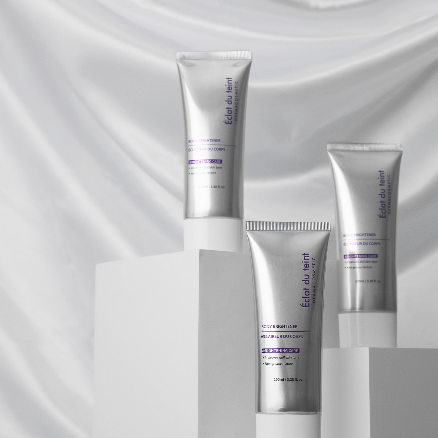 Three silver skincare bottles of Eclat du teint body brightener purple labels on a gray fabric background